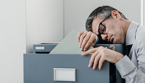 Tired lazy office worker leaning on a filing cabinet and sleeping, he is falling asleep standing up; stress, unproductivity and sleep disorders concept
