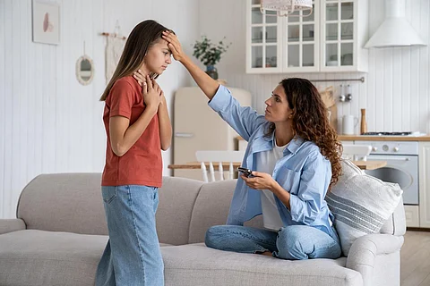 Unhealthy teen girl child complaining of sore throat to mother, mom holding mobile phone touching forehead of schoolgirl checking fever by hand, parent leaving sick kid at home. Influenza in children