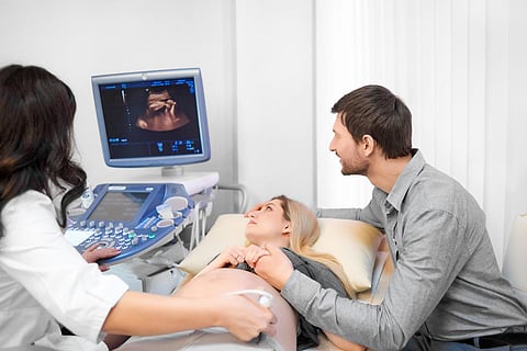Vista trasera de una doctora examinando el estómago de la mujer usando una herramienta especial y un gel. Mamá mirando la foto del bebé. Marido encantador llevando a la esposa embarazada en el procedimiento de ecografía en el hospital.