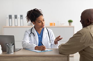 Consultation Concept. Portrait of young black general practitioner doctor talking to male patient during appointment at office, explaining prescription, writing. Therapist sitting with man at desk