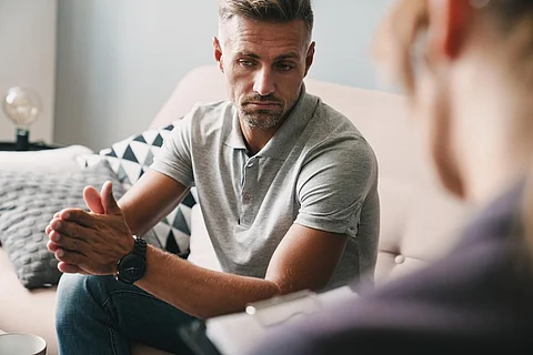 Photo of brooding unshaven man having conversation with psychologist on therapy session in room