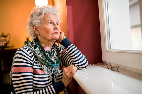 Sad alone senior woman looking through window at home, loneliness concept