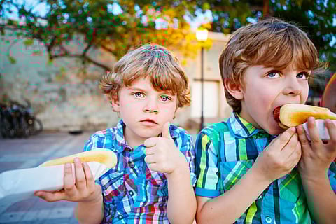 Two little kid boys eating hot dogs outdoors