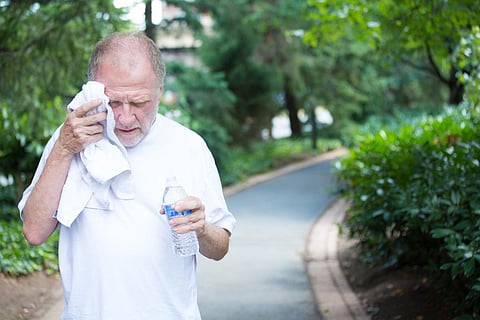 Retrato en primer plano, anciano caballero con camisa blanca que tiene dificultades con el calor extremo, la alta temperatura, se seca el sudor de la cara, muy cansado, aislado fondo de camino pavimentado de árboles verdes