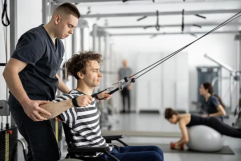 Rehabilitation specialist helps a guy in a wheelchair to do exercise on decompression simulator for recovery from injury. Concept of physical therapy for people with disabilities.