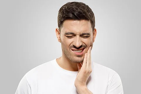 Hombre joven presionando los dedos contra su mandíbula, tratando de superar un fuerte dolor de muelas, con los ojos cerrados, aislado sobre un fondo gris