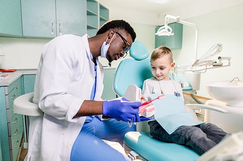 Un dentista negro feliz y sonriente le dice a un niño caucásico cómo cepillarse los dientes. Prevención de caries, odontología, concepto de higiene dental