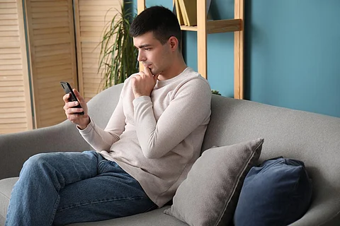 Thoughtful young man with smartphone sitting on sofa