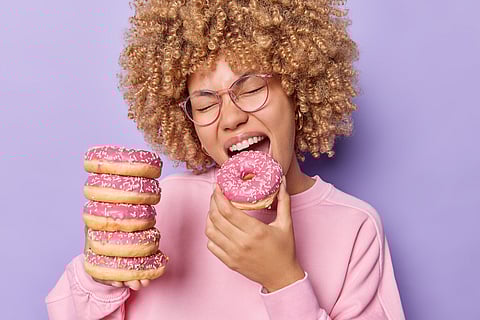 Foto de una mujer golosa hambrienta con el pelo rizado, muerde deliciosa y apetitosa rosquilla glaseada, mantiene la boca bien abierta, disfruta de su comida favorita o merienda, vestida casualmente aislada sobre fondo morado.