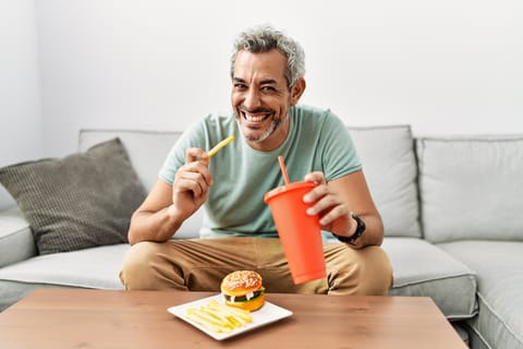 Hombre de pelo gris de mediana edad comiendo patatas fritas sentado en el sofá en casa