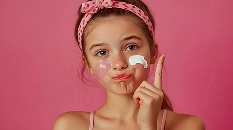 Chica joven con diadema de lunares que aplica crema facial, expresión juguetona sobre fondo rosa. Rutina de cuidado de la piel y concepto de belleza para adolescentes.