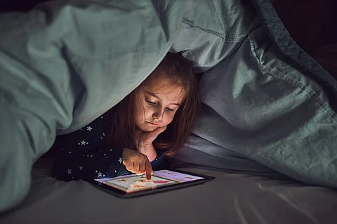 Niña que se divierte viendo, jugando y escuchando cuentos en la tableta. Niño divirtiéndose antes de acostarse acostado debajo del edredón usando la tableta