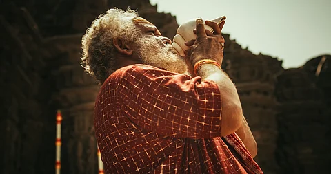 Hinduismo, religión del sur de Asia y metraje cultural: Foto auténtica de un monje indio senior soplando una caracola en un antiguo templo de piedra. Anciano realizando un ritual sagrado en un día de adoración