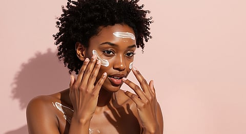 Joven negra aplicándose crema facial durante su rutina de cuidado de la piel. Retrato de modelo afroamericana con piel radiante usando producto cosmético.
