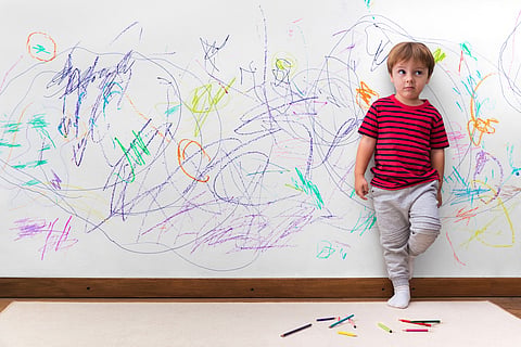 Travesuras infantiles. Chico con cara distraída porque dibujó toda la pared. Niño pequeño apoyado en la pared blanca donde hizo muchos dibujos con lápices de colores. Niño dentro, en casa