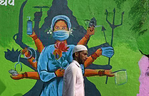The first two waves of the Covid-19 pandemic possibly claimed four million lives in India. Still, the question of healthcare remains absent from national politics.
A man walks past a mural showing a health worker and a shadow of a Hindu deity, painted to create awareness about the spread of Covid-19 in Mumbai. November 2021. Photo: ZUMA Wire / IMAGO