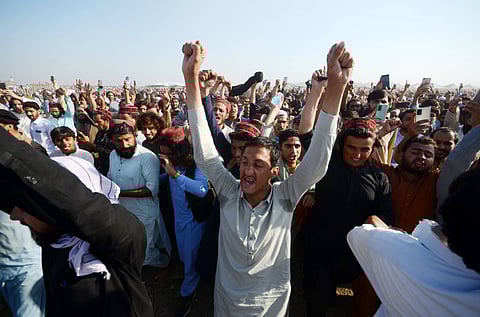 A large crowd of Pashtun men dressed in their traditional attire of kurta and salwar raise their hands as they appear to cheer or chant slogans in an open field