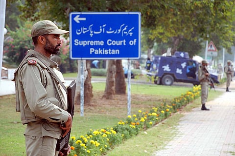 A guard in a khaki uniform and holding a rifle stands in profile in front of a blue sign with white writing pointing towards the Supreme Court of Pakistan