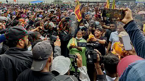 A large crowd of Aragalaya protesters gather around two individuals breaking coconuts. Several activists at the scene are holding phones and cameras to record the event.