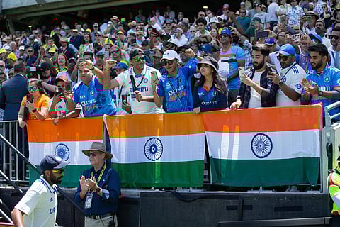 A crowd of Indian cricket fans, many in blue jerseys, behind a row of three Indian flags in the stands during the first Test of the 2024–25 Border-Gavaskar series with Jasprit Bumrah, in cricket whites and wearing the blue India cap, in the foreground.