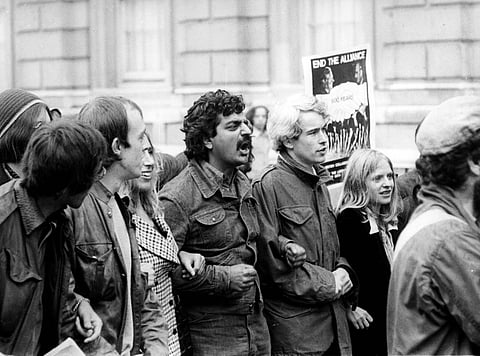 A black and white image of a group of protesters walking side by side with arms interlinked. One protester is carrying a poster that reads "end the alliance".