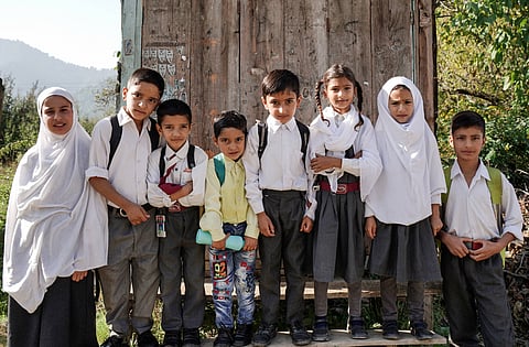 A group of eight Kashmiri boys and girls, dressed in school uniforms and carrying schoolbags and water bottle, pose for a photo in a hilly village in Anantnag district.