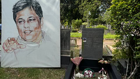Photo of a gravesite in Sri Lanka. The gravestone is that of the murdered Sri Lankan editor Lasantha Wickrematunge, and there is a small portrait of Lasantha Wickrematunge on a blue background embedded in the gravestone. The grave has flower bouquets and candles on it. On the left hand side is a large hand-drawn portrait of Lasantha Wickrematunge, with his left hand out as he explains something. He is smiling.