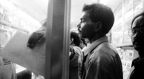A black-and-white photograph captures a group of migrants in a crowded post office in Dubai. A man holding a letter is in focus. Behind him, several other individuals are partially visible, including a man wearing glasses on the right side of the frame.