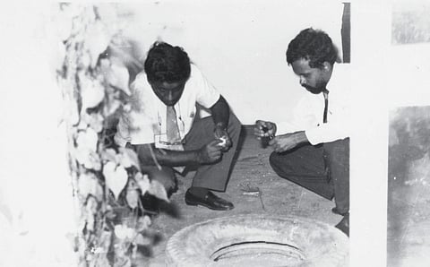 An old black and white photograph of two men in white shirts and ties crouching on the ground near a tyre. The man on the right is holding a small container while the other on the left appears to be handling an object. The setting is a semi-enclosed space, with a white wall in the background and a leafy vine partially obscuring the left side of the frame.
