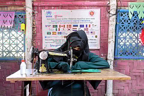 A Rohingya woman dressed in an abaya and hijab works at a sewing machine in a. refugee camp. A banner with logos of donor organisations hangs in the background.