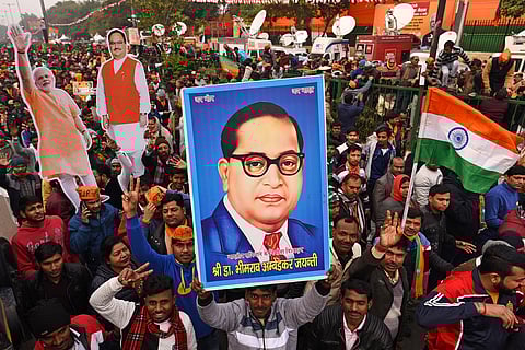 A large crowd of people participates in a political rally. Many in the crowd are holding up signs, including a large portrait of B R Ambedkar. There are also cutouts of Narendra Modi and J P Nadda. The Indian national flag is being waved, and several participants are wearing scarves and caps with BJP insignia.