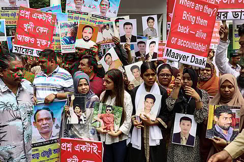 A group of activists in Bangladesh participate in a protest holding photographs of missing persons. They are carrying signs with demands for justice and accountability, one of which reads in English, 'Where are they? Bring them back — alive or accountable! Maayer Daak.' Another sign in Bengali asks why the killers are still free. The protest appears to be organised by families of victims who have killed or disappeared, likely in cases of enforced disappearances.
