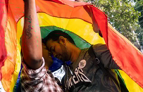 Two men stand face to face under a rainbow pride flag at a Pride Parade in Guwahati, Assam. One of them wears a plaid shirt with a visible tattoo on their arm, while the other wears a dark shirt with a button reading “Queer Pride.”