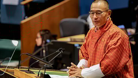 Tshering Tobgay in traditional Bhutanese attire, wearing glasses and a red patterned gho, stands at a podium delivering a speech at the United Nations General Assembly in September 2024. The background setting is an assembly hall, with the United Nations emblem visible on the front of the podium. Multiple microphones and papers are in front of him. Blurred attendees and tiered seating are also visible in the background.