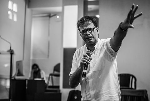 A black-and-white photo of Jerry Pinto in a white shirt holding a microphone and gesturing emphatically with his arm extended forward. He is speaking to an audience in an indoor setting, with blurred figures and furniture visible in the background.
