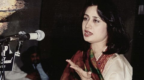 A photograph of Parveen Shakir in speaking into a microphone at a public event. She is mid-sentence, with one hand raised in an expressive gesture. She's wearing a cream-coloured kameez with a red and green dupatta and a pearl necklace, and she's sitting on a platform in front of a dark background with another person slightly visible in the distance.