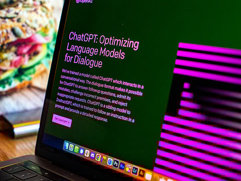 A close-up photo of a laptop screen displaying the ChatGPT website by OpenAI. The screen shows the headline “ChatGPT: Optimizing Language Models for Dialogue” on a dark background, along with a brief description of the model’s purpose. The right side features a stylised pink and black abstract design of its logo.