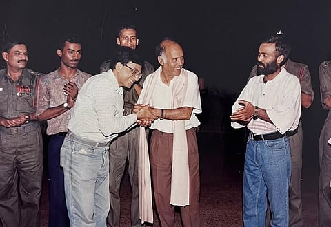 A group of men gathered at a theatre event. At the centre, Swadesh Deepak, wearing a white shawl and brown trousers, smiles warmly while shaking hands with a man in a striped shirt and jeans. Another man in a white shirt and blue jeans stands nearby, applauding. A few actors in military uniforms and casual clothes stand in the background, also clapping.