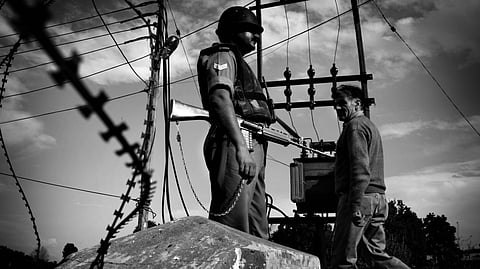 A black-and-white image of a Kashmiri man walking past an armed Indian security personnel standing on a concrete block beside barbed wire, with power lines and a cloudy sky in the background, Srinagar, 2009.