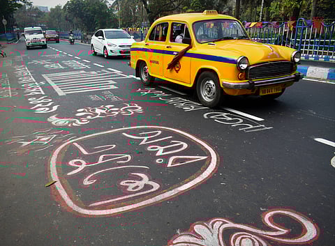 A yellow taxi drives along a street in Kolkata decorated with colourful Bengali messages painted on the asphalt for International Mother Language Day 2025. The messages are written in white and red paint, surrounded by other road markings. Other vehicles are visible in the background, and the roadside is lined with trees and a blue railing adorned with colourful flags.