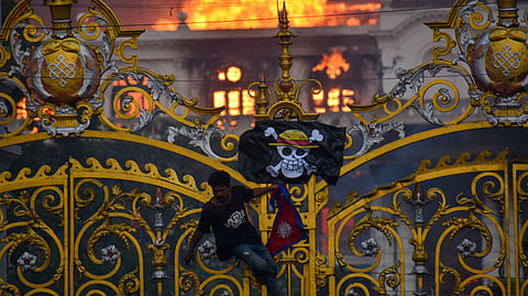 A man hanging from an ornate gate with a pirate flag hung on it as a white neo-classical building burns in the background.