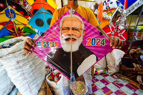 A vendor holds a colourful kite featuring a large image of the Indian prime minister Narendra Modi, with the words "MISSION 2024" printed on either side of his portrait. The kite has a pink and magenta background, and is displayed in a vibrant kite shop filled with other brightly coloured kites.