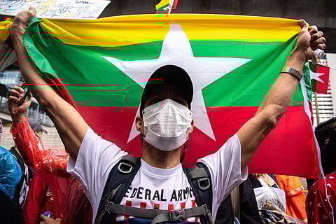 A protester wearing a face mask and cap holds up the flag of Myanmar during a Spring Revolution demonstration in Bangkok. The crowd around him is dressed in colourful clothing, with protest signs visible in the background.