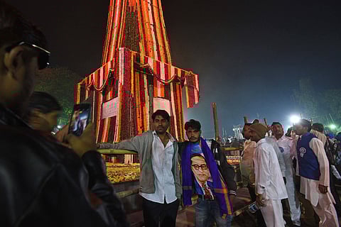 People gather at night around the obelisk commemorating the Battle of Bhima Koregaon, decorated with colourful garlands. Two men pose for a photo in front of the monument while another person takes their picture. One of the men wears clothing featuring an image of B R Ambedkar.