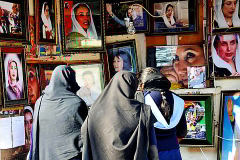 A group of women, seen from behind and wearing dark headscarves and shawls, stand closely together in front of a display wall covered with framed portraits of Benazir Bhutto.