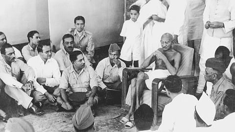 Mohandas Gandhi sits on a chair during a visit to a bhangi colony. He is surrounded by army officers in uniform. Jawaharlal Nehru is visible on the right, and Vallabhai Patel is visible in the middle.