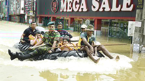 Photo shows a rubber dinghy moving through high floodwater. A man in Sri Lanka Army uniform is in front, along with a volunteer in civilian clothes. There are two more male civilian volunteers in the back. The dinghy has a woman, child and man being rescued from floods. All three wear orange lifejackets. This is in Wellampitiya, Sri Lanka