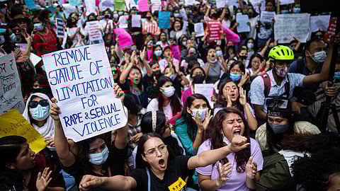 A large crowd of protesters in Kathmandu chant and raise fists while holding signs during a demonstration. In the foreground, a woman holds a handwritten sign reading “Remove statute of limitation for rape & sexual violence”.
