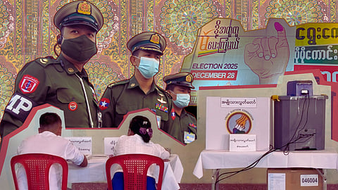 Composite image of Myanmar military junta looming over an electronic voting machine with election posters on the top right, and a stained glass pattern in the background.