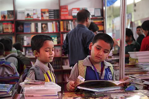 Two young schoolboys with backpacks stand at a bookstall, flipping through illustrated books, while other children and adults browse shelves filled with books in the background.
